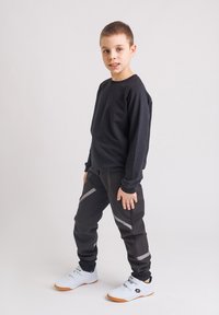 Chłopiec stoi ubrany w czarną bluzę, czarne spodnie z szarymi paskami i białe trampki na jednolitym jasnym tle.