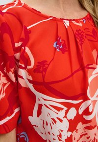 Rote Bluse mit rundem Ausschnitt, ausgestattet mit einem auffälligen Blumenmuster in Rottönen und Blau, aus weichem, leicht strukturierten Stoff.