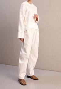 Weißer Pullover mit geripptem Design, kombiniert mit lockeren weißen Hosen und braunen Stiefeletten mit Schlangenmuster.