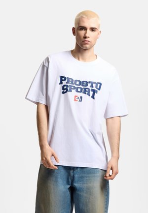 Młody mężczyzna z krótkimi blond włosami, ubrany w białą koszulkę "PROSTO SPORT" i luźne niebieskie jeansy, stojący na jednolitym tle.