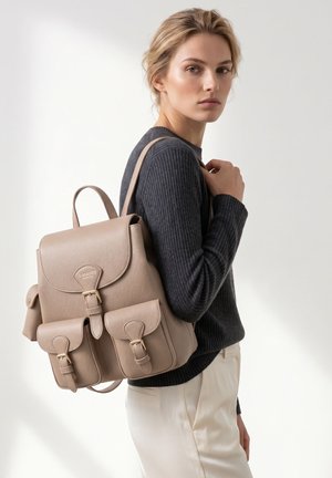 Vrouw die een zwart geribde trui en crèmekleurige broek draagt, heeft een beige leren rugzak met voorvakken met gesp over één schouder hangen.