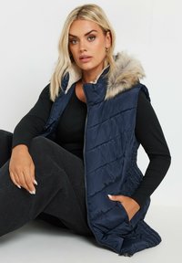 Laivastonsininen puffertakki, jossa on tekoturkiskaulus, tikattu muotoilu, vetoketjukiinnitys ja sivutaskut. Päällä musta pitkähihaisen paita.