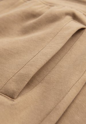 Detail béžové látky zobrazující vertikální otevření kapsy s čistým šitím na měkkém texturovaném materiálu.