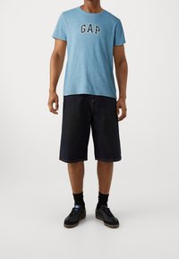 Blaue Baumwoll-T-Shirt mit "GAP"-Logo, kombiniert mit dunklen Jeans-Shorts bis zum Knie und schwarzen Sneakers mit blauen Akzenten.