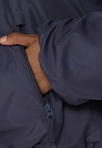 Hand in der mit Reißverschluss versehenen Tasche einer dunkelblauen Jacke mit elastischen Bündchen und dezentem Logo auf dem Ärmel.