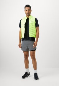 Chaleco acolchado verde lima con cremallera en el frente, usado sobre una camiseta negra. Combinado con pantalones cortos grises y zapatillas deportivas negras.