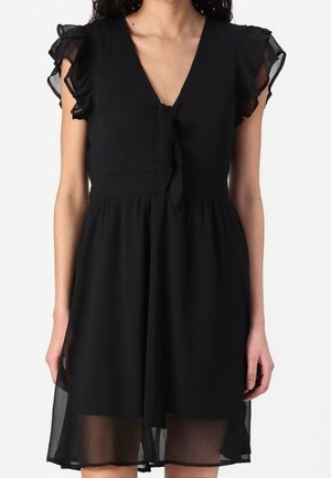 Robe noire sans manches avec des manches courtes volantées transparentes et un nœud noué en V à l'avant, portée par une femme debout.
