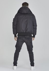 Schwarze Daunenjacke mit Kapuze und gerippten Bündchen, mit einem auffälligen Logo am Kragen. Tragen mit dunklen Cargo-Hosen und weißen Sneakers.