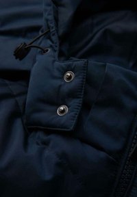 Giacca isolamento blu navy con una texture liscia, caratterizzata da bottoni a pressione metallici e una chiusura con cordino al colletto per una vestibilità regolabile.