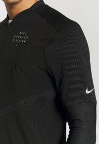 Close-up van een persoon die een zwarte Nike Running Division jas draagt met geribbelde mouwen en een witte Nike swoosh op de bovenarm.