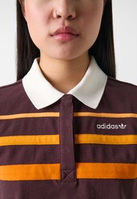 Izbliza osoba u tamnoplavoj polo majici s bijelim ovratnikom i narančastim horizontalnim prugama, koja prikazuje Adidasov logo na prsima.