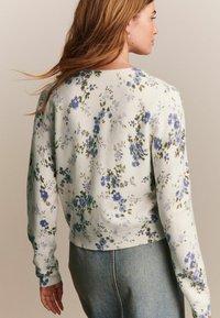 Pull en fleurs avec un fond crème, orné de fleurs bleues et vertes. Manches et ourlet côtelés, design court. Texture en tricot douce.