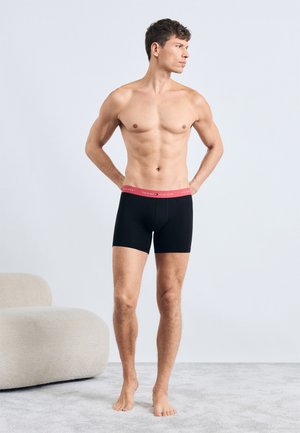 Sorte boxershorts med en pink elastik, der har brandets logo, båret af en mandlig model med siden til, barfodet på et lyst tæppe.