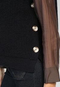 Gilet en mailles bleu marine avec de grands boutons crème sur le côté, associé à une manche en tissu transparent marron. Tissu texturé et détail de bord-côte.