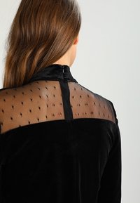 Schwarzes Samtoberteil mit transparenter Netzrückseite, das ein gepunktetes Muster aufweist. Hoher Kragen und Reißverschlussverschluss hinten. Weiche Textur.