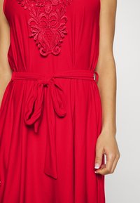 Vestido vermelho com bordados intrincados na zona do decote, um cinto a condizer atado na cintura e uma textura de tecido fluido.