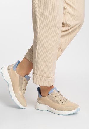 Beige Sportschuhe mit hellblauen Akzenten, aus Wildleder und Leder, mit strukturierten Einsätzen und flexibler weißer Sohle. Die Hose ist aus beigem Cord.