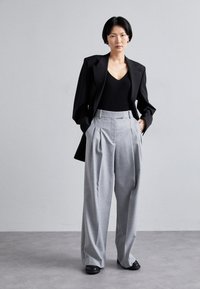 Black tailored blazer over a ribbed black top, paired with light gray pleated wide-leg trousers and black shoes, against a neutral background.