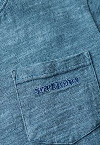 Blå struktureret bomulds skjorte med en brystlomme, hvor "SUPERDRY" er broderet med mørkere blå tråd. Casual design med synlige syninger.