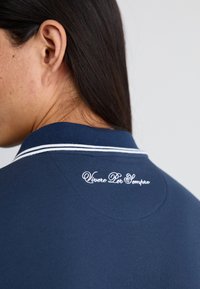 Marinblå polo skjorte med ribbestrikket krage og hvite detaljer, med dekorativt brodert tekst "Vivere Per Sempre" på baksiden.