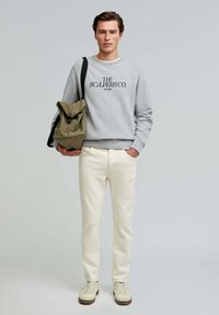 Gray sweatshirt with navy text, cream trousers, and beige sneakers. Model holds a folded olive backpack with a black strap.
