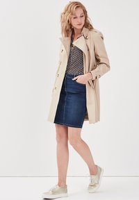Manteau beige avec des boutons dorés, blouse à motifs, jupe en jean sombre et baskets de couleur claire, présentant des tissus texturés et des détails de couture.