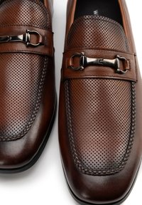 Mocassins en cuir marron avec un design perforé, un accent de boucle en argent et des coutures contrastées le long des bords.