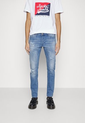 Jeans in denim blu con vestibilità slim, caratterizzati da dettagli consumati e strappi. Abbinati a una maglietta bianca con un logo grafico rosso e blu navy.