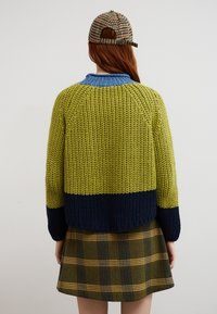 Femme aux longs cheveux roux portant un pull épais tricoté vert, bleu et marine, une jupe à carreaux et une casquette pied-de-poule, tournée dos à la caméra.