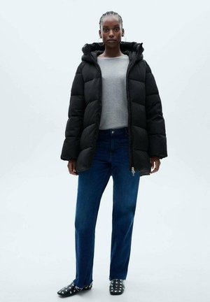 Black puffer jacket with a hood, featuring large quilted sections. Paired with a gray long-sleeve shirt and dark blue jeans. Black shoes with studs.