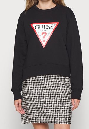 Sudadera negra con triángulo del logo Guess en rojo y blanco, combinada con falda de pata de gallo en blanco y negro, en una persona de pie.