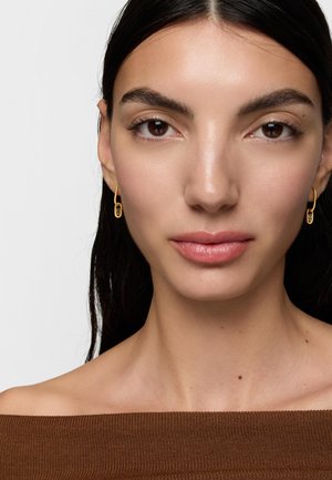 Donna con capelli scuri che indossa orecchini pendenti dorati e un top marrone senza spalline, rivolta in avanti con un'espressione neutra.