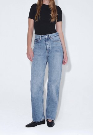 Femme portant un jean bleu clair taille haute droit, un haut noir à manches courtes et des chaussures plates noires sur fond uni.