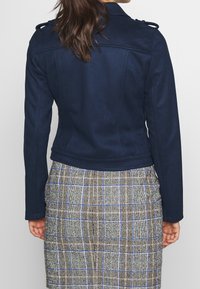 Mörkblå, figursydd jacka med slät textur, axelklaffar och kort längd, kombinerad med en rutig kjol i jordnära toner.