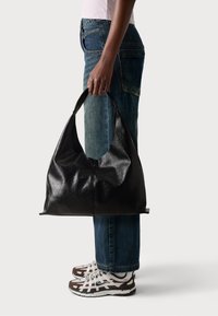 Svart läder hobo-väska med slät yta och böjd handtag, formad för enkel bärning, hålls av en person som bär blå jeans och sneakers.