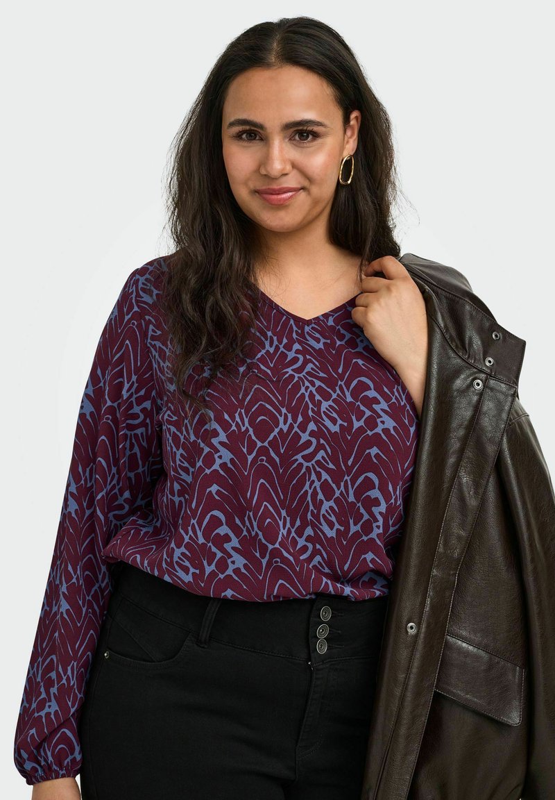 Femme aux longs cheveux foncés porte un chemisier violet à motifs et un pantalon noir, tenant une veste en cuir brun foncé sur son épaule.