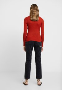 Rode geribde top met lange mouwen, nauwsluitend ontwerp, gecombineerd met zwarte jeans. Zwarte gekruiste sandalen maken de outfit compleet. Witte achtergrond.