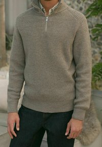 Beige ribbed knit sweater with a quarter-zip collar, paired with dark denim jeans. Warm texture and fitted design, no visible patterns.
