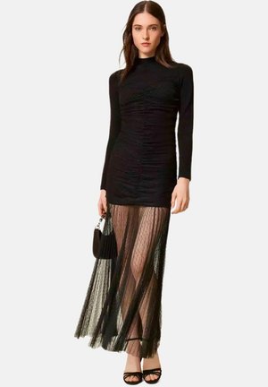 Donna che indossa un mini abito nero a maniche lunghe con una sovrapposizione maxi nera plissettata e trasparente, tiene in mano una piccola borsa nera e indossa tacchi neri con cinturini.