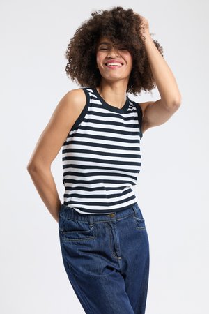 Uśmiechnięta kobieta o kręconych włosach, ubrana w czarno-biały, bezrękawnik w paski oraz luźne niebieskie dżinsy, pozująca z ręką we włosach.