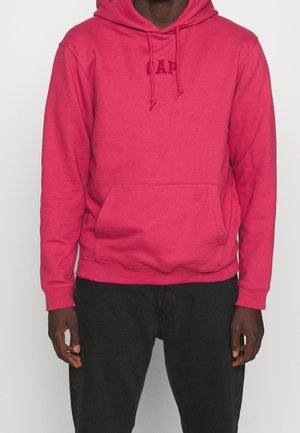 Persoon die een felroze hoodie draagt met een kangoeroezak en bijpassende trekkoorden, gecombineerd met een zwarte broek, staand tegen een witte achtergrond.