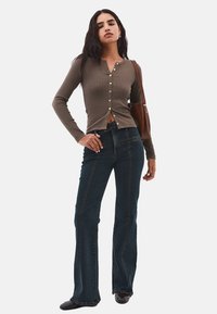 Camisa marrón de manga larga con textura acanalada y cierre de botones, combinada con unos vaqueros de campana azul oscuro. Colores sólidos, textura suave, con un diseño minimalista.