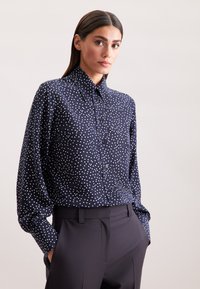 Camicia navy con bottoni, a pois bianchi, con colletto classico e maniche lunghe, abbinata a pantaloni scuri su misura. Tessuto morbido.