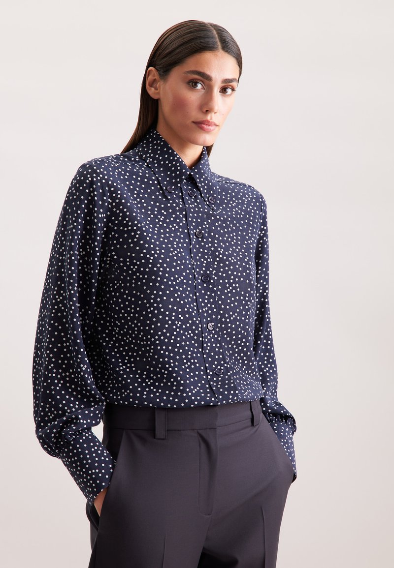 Camicia navy con bottoni, a pois bianchi, con colletto classico e maniche lunghe, abbinata a pantaloni scuri su misura. Tessuto morbido.