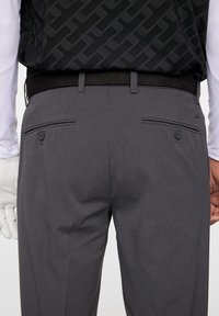 Szare spodnie z subtelnym teksturowanym wzorem, tylne kieszenie z zapięciem na guziki oraz gładki czarny pasek. Widoczna biała rękaw.
