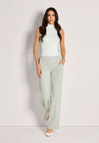 Vrouw met lang donker haar, gekleed in een mouwloze witte ribbed top, lichte geblokte wijde broek en witte puntige hakken, staat zelfverzekerd.