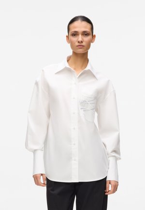 Kobieta ubrana w za dużą białą koszulę zapinaną na guziki, z bufkami na rękawach i sznurowanym detalem na kieszeni na piersi, w połączeniu z czarnymi spodniami.