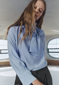Blusa azzurra con un dettaglio di sciarpa annodata al collo e maniche lunghe. Tessuto liscio, abbinata a pantaloni a quadri scuri.