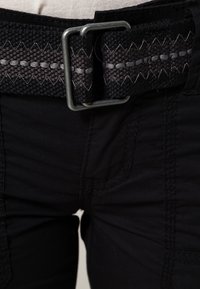 Ceinture tissée noire avec un motif zigzag gris, dotée d'une boucle en métal. Portée par-dessus un pantalon en tissu noir avec des détails de couture visibles.