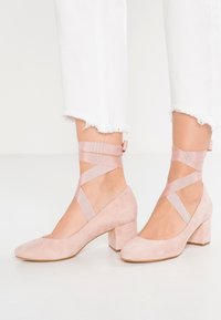 Růžové suede boty na blokovém podpatku s pásky kolem kotníku. Kulatá špička a strukturovaný povrch. Nosené s bílými džíny s roztrženými lemy.